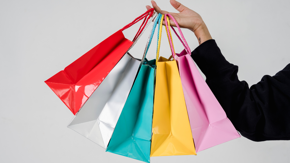 Hand holding colorful red, silver, blue, yellow, and pink shopping bags against a light background.