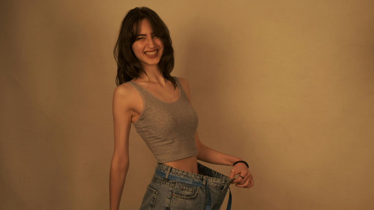 Woman smiling while holding the loose waistband of oversized jeans, representing weight loss and body transformation.